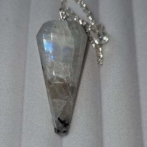 May include: A faceted, clear crystal pendulum with a silver chain. The crystal has a pointed bottom and a geometric shape. The crystal has a rainbow sheen and some dark inclusions. The chain is attached to the top of the crystal.