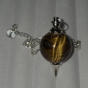 May include: A pendulum with a silver chain and accents. The pendulum features a spherical, tiger's eye stone with brown and gold striations. The pendulum has a silver tip and a silver cap at the top.