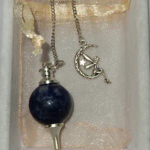 May include: A blue lapis lazuli crystal pendulum with a silver chain and a silver charm of a fairy sitting on a crescent moon.