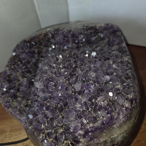 May include: A large, raw amethyst geode with a deep purple hue. The crystal formation is visible, with many small, sparkling points. The geode sits on a wooden surface, and a black cord is visible.