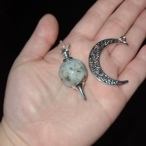 May include: A silver pendulum with a clear crystal ball and a silver crescent moon charm. The pendulum is hanging from a silver chain.