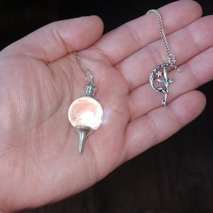 May include: A silver pendulum with a clear, pink-tinted crystal ball and a silver chain. The pendulum is hanging from a hand. A second silver charm with a crescent moon and a fairy design is also hanging from a chain.