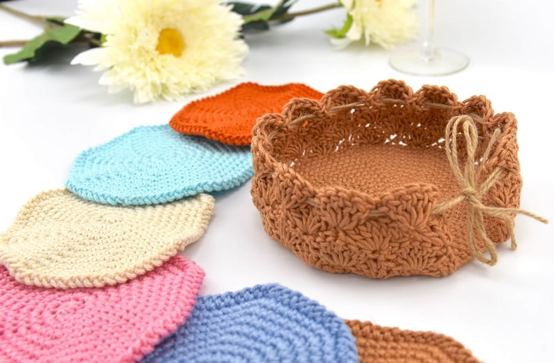 May include: A set of crocheted coasters in various colors, including orange, blue, cream, pink, and light blue, with a larger crocheted basket in a brown color.