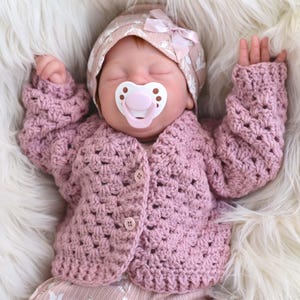 May include: A baby wearing a pink crocheted cardigan and matching hat with a bow, and a pacifier. The baby is lying on a white fluffy surface. The cardigan has three buttons.