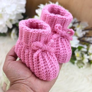 May include: A pair of handmade pink crochet baby booties. The booties feature a ribbed cuff and a small bow detail on the front. The soft pink color and delicate design suggest a newborn or infant item.