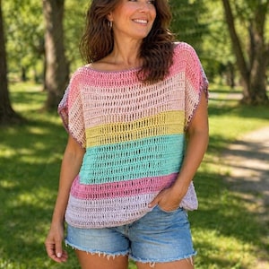 May include: A colorful, striped crochet top in shades of pink, yellow, green, and lavender. The top has a loose, boxy fit with short sleeves. The model is wearing denim shorts. The background is a park with trees.