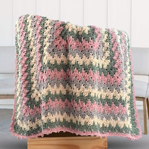 Puede incluir: Una manta de ganchillo en tonos rosa, verde y beige con un patrón de chevron. La manta está doblada y colocada sobre una caja de madera.
