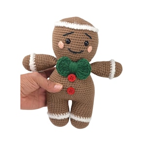 May include: A brown crocheted gingerbread man toy with a green bow tie and red buttons. The toy has a white icing trim around the head and feet.