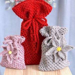 Op de afbeelding: Drie handgemaakte gehaakte cadeauzakjes in rood, lichtpaars en grijs. De rode tas is lang en vastgebonden met een trekkoord. De lichtpaarse en grijze tassen hebben een bloemenapplicatie met een gele kern. De tassen staan op een houten oppervlak.