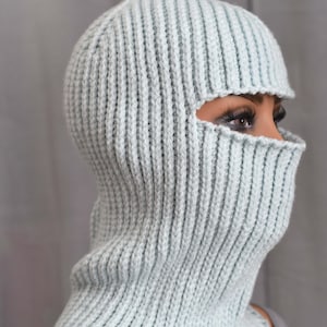 Könnte beinhalten: Hellblau gestrickte Sturmhaube mit Rippstruktur. Die Kopfbedeckung hat Öffnungen für Augen und Mund sowie einen Nackenwärmer. Die Sturmhaube ist aus einem weichen, warmen Material gefertigt.