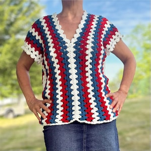 May include: A crochet top in a patriotic color scheme of red, white, and blue. The top features a V-neck and short sleeves with a scalloped edge. The crochet pattern creates vertical stripes.