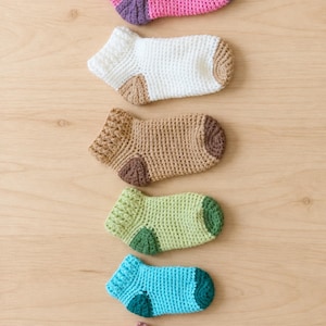 May include: A vertical arrangement of six ankle socks crocheted in various colours including pink, white, tan, light green, bright blue, and dusty rose. Each sock features a textured cuff, a solid main body, and a contrasting colour on the heel and toe.