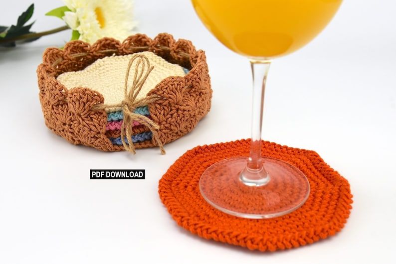 May include: A set of crocheted coasters in a variety of colors, including beige, pink, blue, and orange. The coasters are stacked in a basket made of the same crocheted material. A glass of orange juice sits on top of one of the coasters.