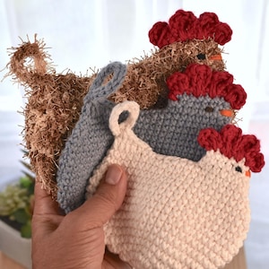 May include: A hand holds a stack of handmade crochet chicken-shaped pot holders in various colors. The pot holders are beige, gray, and brown, with red comb details. Each has a loop for hanging.