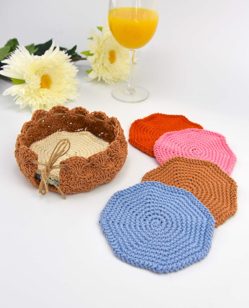 May include: Set of five crocheted coasters in various colors, including orange, pink, brown, and blue. The coasters are arranged in a circle around a crocheted basket.