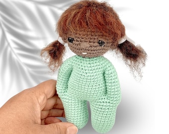 May include: A crocheted doll with brown hair, brown skin, and a light green outfit. The doll has a sweet expression and is wearing white bows in her hair.