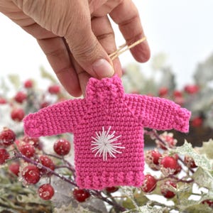 May include: A pink crocheted sweater ornament with a white snowflake design. The ornament is hanging from a twine string.