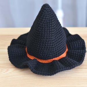 May include: A black crochet witch hat with a pointed crown and a wide, ruffled brim. An orange band encircles the base of the cone-shaped crown. The hat is displayed on a light-colored wooden surface.