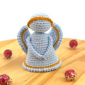 Pode incluir: Um ornamento de anjo de crochê com fio azul e uma auréola amarela. O anjo está em pé sobre uma superfície de madeira.