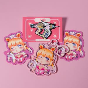 May include: A collection of pink and white Juno-themed merchandise, including a pin, keychains, and stickers. The pin features a colorful ray gun design with the name "Juno" at the top. The stickers and keychains depict a cartoon girl blowing a kiss.