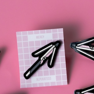 May include: Four black and pink enamel pins shaped like a closed umbrella. The pins are on a pink background. One pin is on a white card with the text "MERCY" and "ALPUKATSSS".
