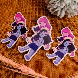 May include: Three sticker designs featuring cartoon characters. The stickers are printed on white paper and feature a pink-haired character sitting on a blue-haired character. The pink-haired character is holding a book in one design and a video game controller in the other two designs.