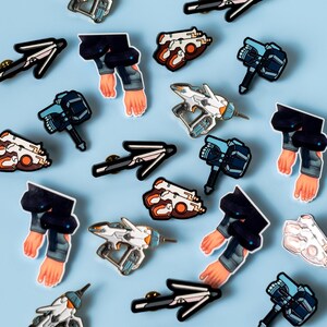 May include: A collection of enamel pins featuring various designs, including spaceships, weapons, and human arms. The pins are on a light blue background.