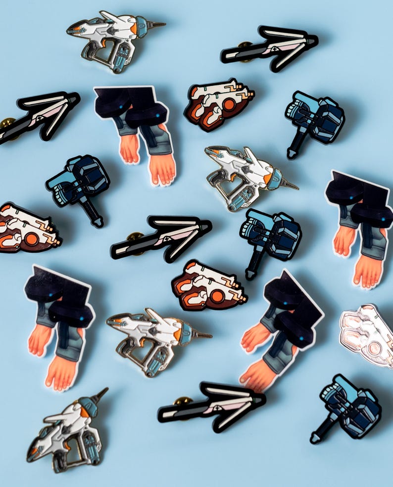 May include: A collection of enamel pins featuring various designs, including spaceships, weapons, and human arms. The pins are on a light blue background.