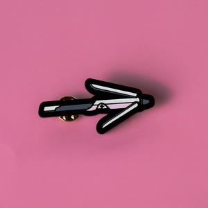 May include: Black and white enamel pin shaped like a hair straightener, with a pink stripe on the top. The pin has a gold back and a safety clasp.