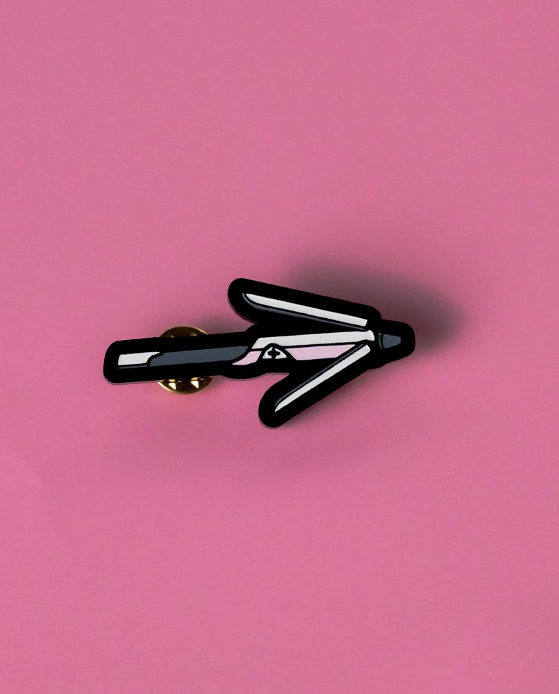 May include: Black and white enamel pin shaped like a hair straightener, with a pink stripe on the top. The pin has a gold back and a safety clasp.