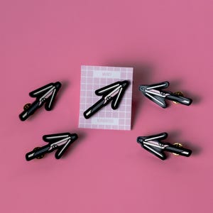 May include: Five black and pink enamel pins shaped like an arrow pointing to the right. The pins are on a pink background. A white card with a grid pattern and the words "Mercy" and "Alphonsosss" is in the background.