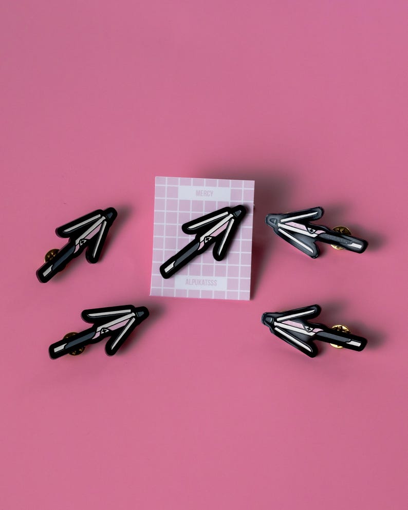 May include: Five black and pink enamel pins shaped like an arrow pointing to the right. The pins are on a pink background. A white card with a grid pattern and the words "Mercy" and "Alphonsosss" is in the background.