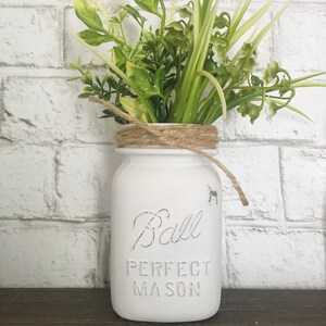 May include: White painted Ball Perfect Mason jar with a brown twine bow and a bouquet of green leafy plants.