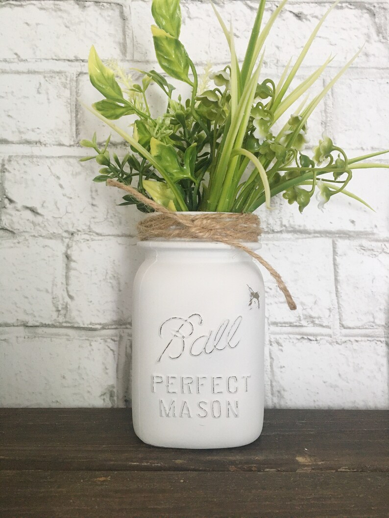 May include: White painted Ball Perfect Mason jar with a brown twine bow and a bouquet of green leafy plants.