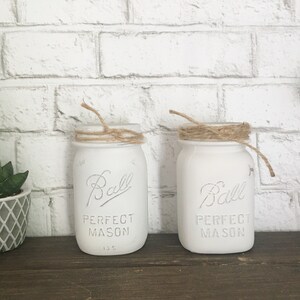 May include: Two white painted Ball mason jars with twine tied around the top. The jars are labeled "Ball Perfect Mason" and have the number "135" printed on the bottom.