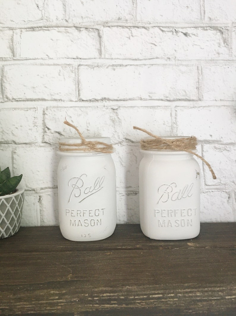 May include: Two white painted Ball mason jars with twine tied around the top. The jars are labeled "Ball Perfect Mason" and have the number "135" printed on the bottom.