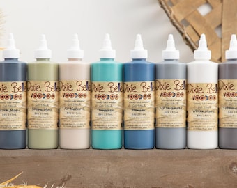 May include: Eight bottles of Dixie Belle Voodoo Gel Stain in various colors, including black, green, beige, teal, blue, gray, white, and brown. Each bottle is labeled with the Dixie Belle Voodoo logo and the name of the color.