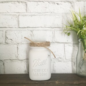 May include: White painted Ball Perfect Mason jar with a brown twine bow around the lid. The jar is sitting on a brown wooden surface.