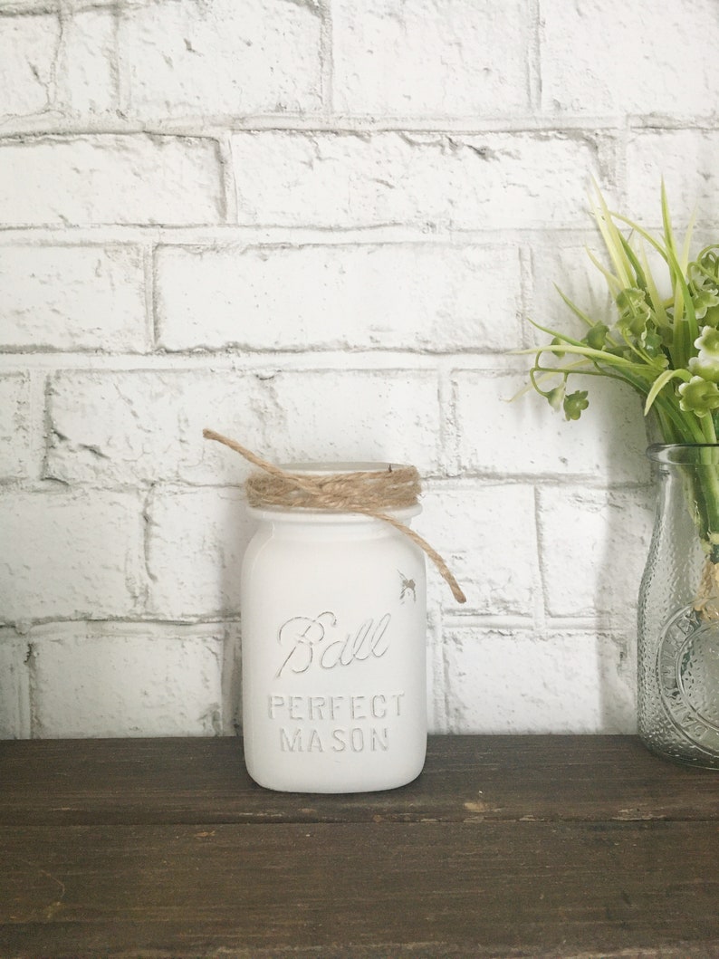 May include: White painted Ball Perfect Mason jar with a brown twine bow around the lid. The jar is sitting on a brown wooden surface.