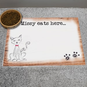 May include: A white wooden pet feeding mat with the text "Missy eats here..." and a drawing of a cat wearing a collar. The mat has a brown wood grain pattern and two black paw prints.