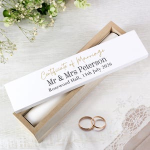 May include: A wooden box with a white lid, inscribed with "Certificate of Marriage", names, date, and location in gold lettering. Two gold wedding rings are placed on a white cloth. The box is open.