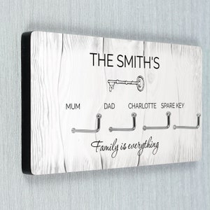 May include: White wooden key holder with black trim. It has four hooks labelled "Mum", "Dad", "Charlotte", and "Spare Key". The text "THE SMITH'S" and a key graphic are at the top. The text "Family is everything" is at the bottom.