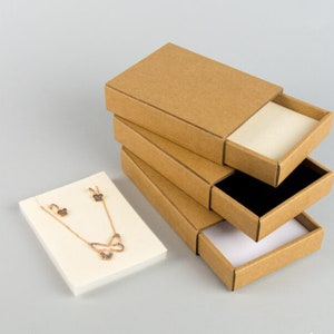 May include: Four brown cardboard boxes with sliding lids, stacked on top of each other. The top box is open, revealing a white interior. A small white square box with a gold necklace and earrings with butterfly designs is in the foreground.