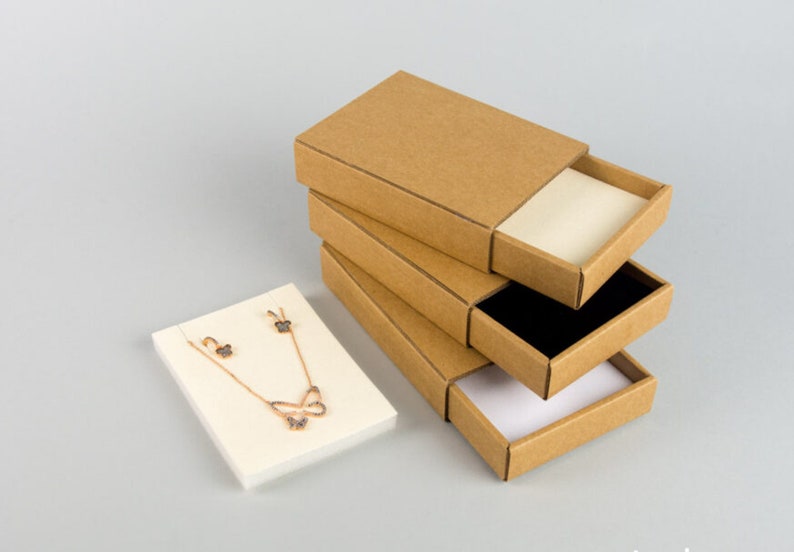 May include: Four brown cardboard boxes with sliding lids, stacked on top of each other. The top box is open, revealing a white interior. A small white square box with a gold necklace and earrings with butterfly designs is in the foreground.