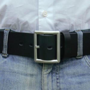 Puede incluir: Un cinturón de cuero negro con una hebilla plateada. El cinturón se lleva alrededor de la cintura de una persona que lleva pantalones vaqueros azules.