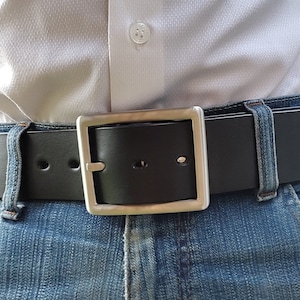 Op de afbeelding: Een zwarte leren riem met een zilveren gesp. De riem wordt gedragen om de taille van een persoon die een blauwe jeans draagt.