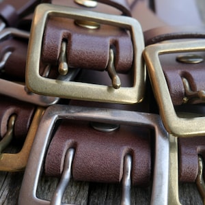 May include: Close-up of multiple brown leather belts with brass and silver buckles. The buckles have a double prong design.