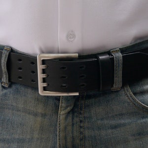 Może przedstawiać: Czarny skórzany pasek ze srebrną klamrą i perforowanym wzorem. Pasek jest noszony na niebieskich dżinsach.