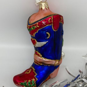 Op de afbeelding: Een blauwe en rode glazen kerstversiering in de vorm van een cowboylaars. De laars heeft gouden accenten en een roze band rond de bovenkant. De versiering heeft een label met de tekst "Mac".