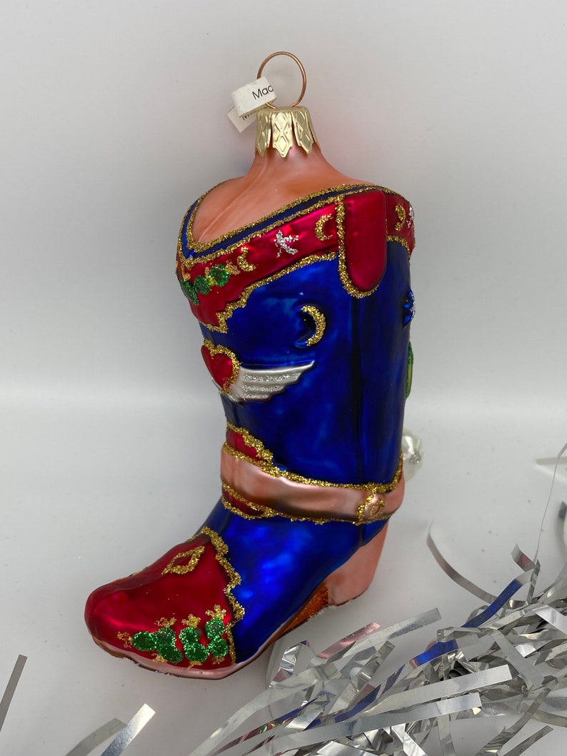 Op de afbeelding: Een blauwe en rode glazen kerstversiering in de vorm van een cowboylaars. De laars heeft gouden accenten en een roze band rond de bovenkant. De versiering heeft een label met de tekst "Mac".